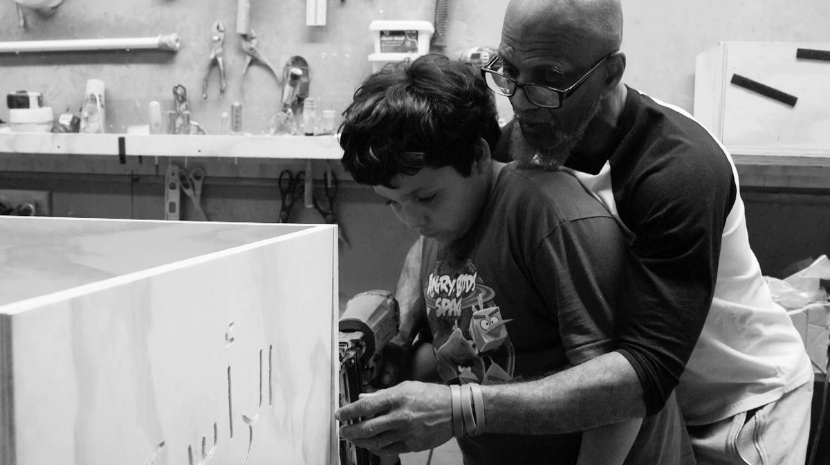 Black and white photograph of a man with dark skin tone helping a young boy use a drill while making a coffin.