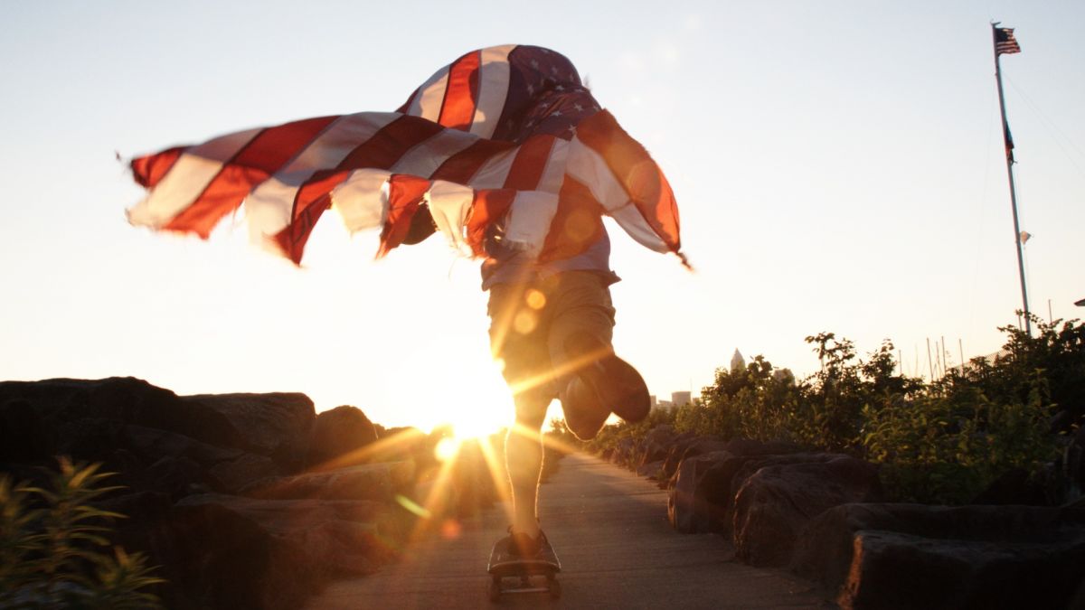 A skateboarder rides with an American flag flying behind them. A lens flare obscures the image slightly.