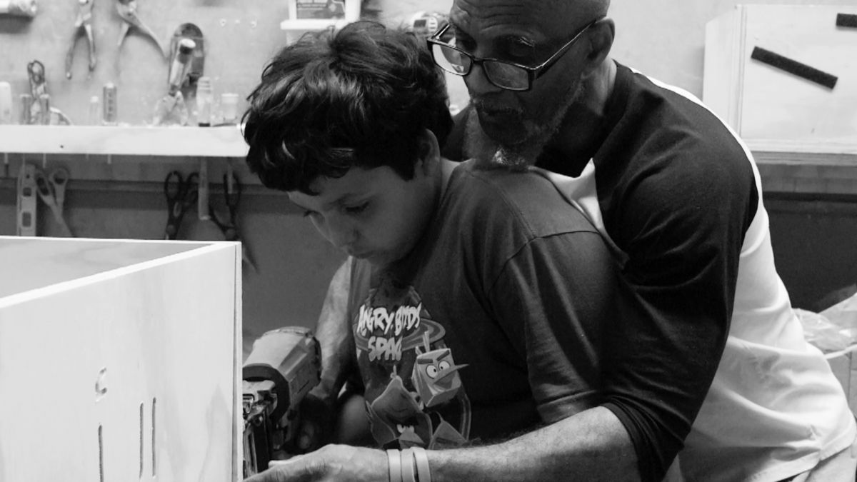 Black and white photograph of a man with dark skin tone helping a young boy use a drill while making a coffin.