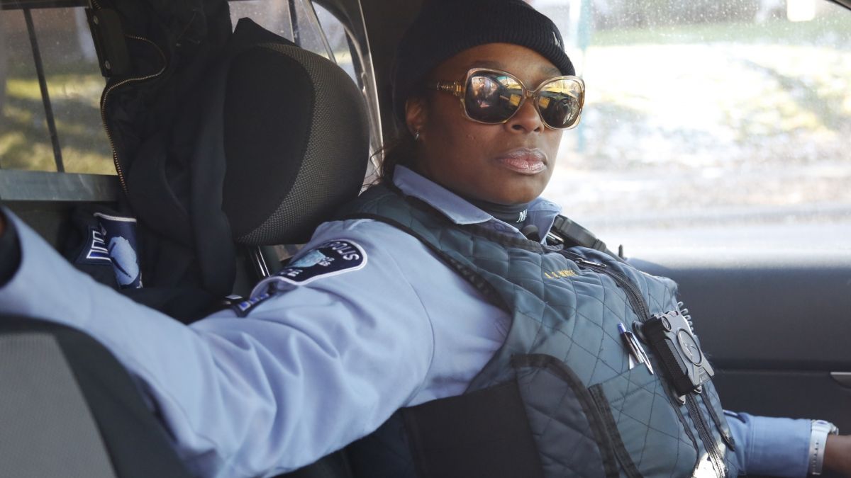 A female police officer, wearing a bullet-proof vest, a blue shirt, and sunglasses, poses in a patrol car with her arm on the passenger seat.