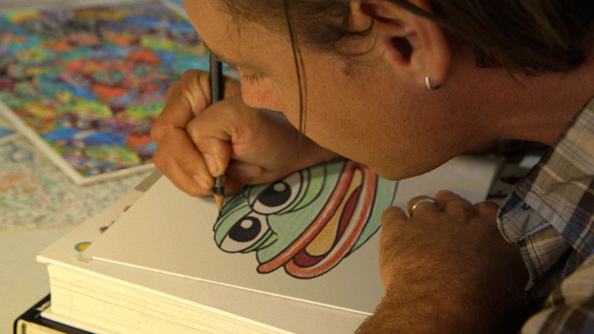 Illustrator Matt Furie, a man with light skin tone, is bent over a table drawing an image of Pepe the Frog.