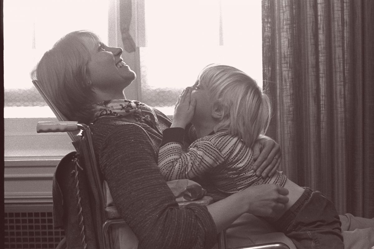 Black and white photograph of a smiling woman using a wheelchair while her young son plays peek-a-boo in her lap.