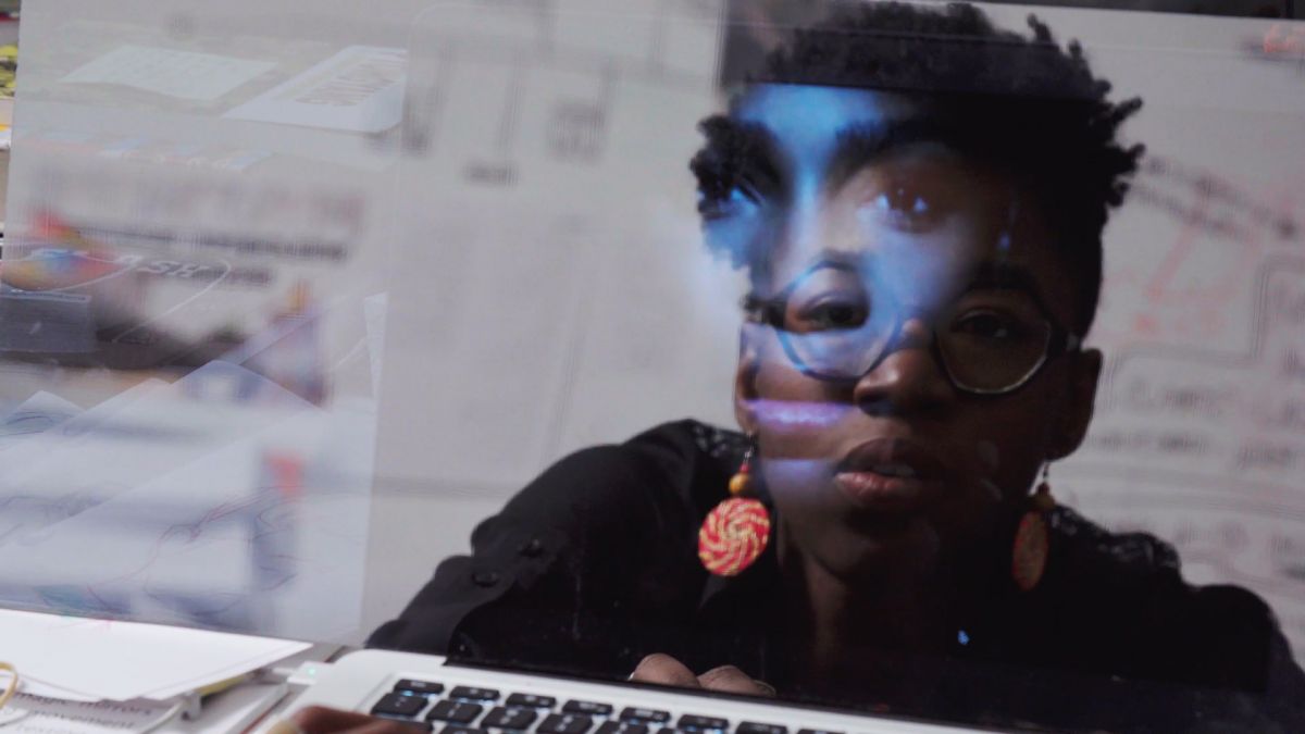 A woman with dark skin tone, short hair, and round, black-rimmed glasses works on a Mac laptop computer. On the laptop screen is her reflection as well as an image of another woman with dark skin tone.