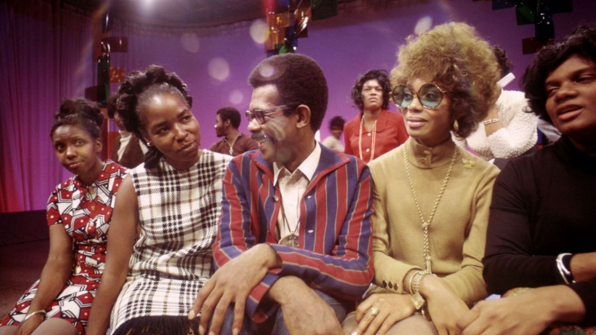 5 people with various shades of skin tone sit together on the set of Soul! They are all wearing fabulous 1960s/70s outfits.