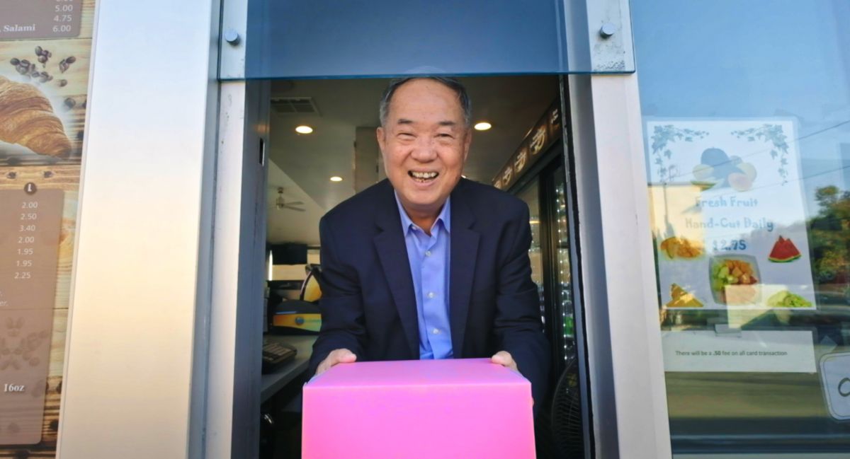 A smiling Cambodian-American man in a suit offers a pink box of donuts through a drive-thru window.