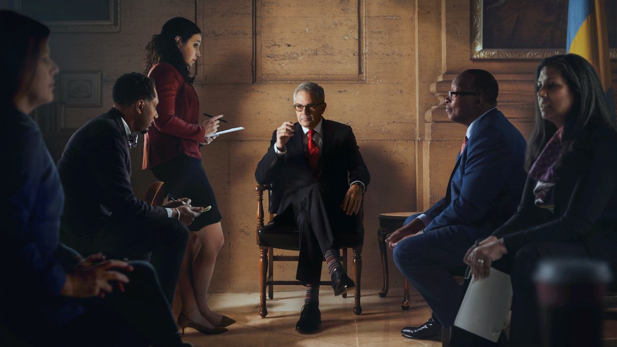 The Philadelphia D.A., a man with light skin tone, gray hair, and glasses, sits in a leather chair flanked by his team: two rows of people with various skin tones wearing business casual attire.