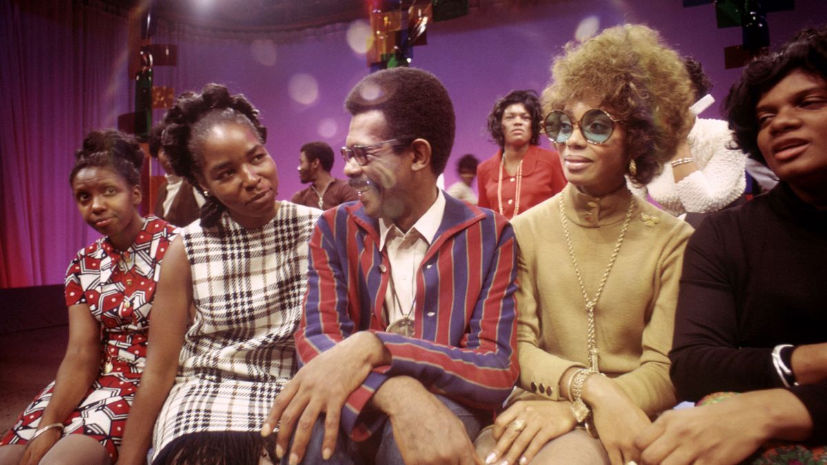 5 people with various shades of skin tone sit together on the set of Soul! They are all wearing fabulous 1960s/70s outfits.