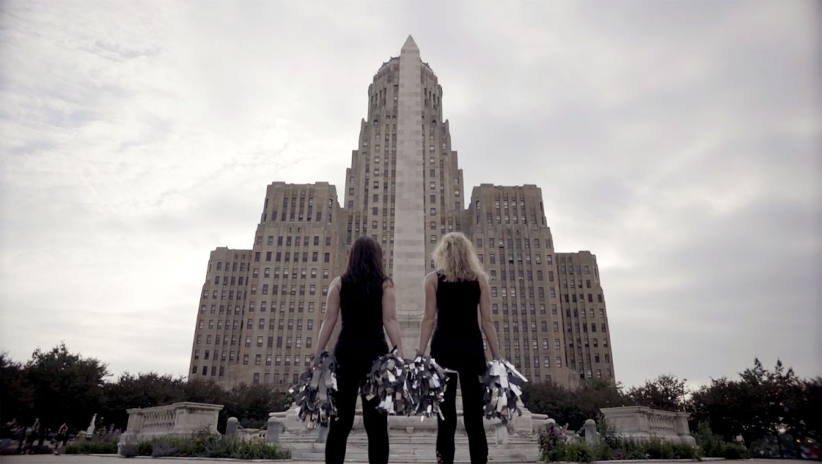 Two NFL dancers with light skin tone pose with their backs to the camera and pom-poms in hand in front of a large courthouse.