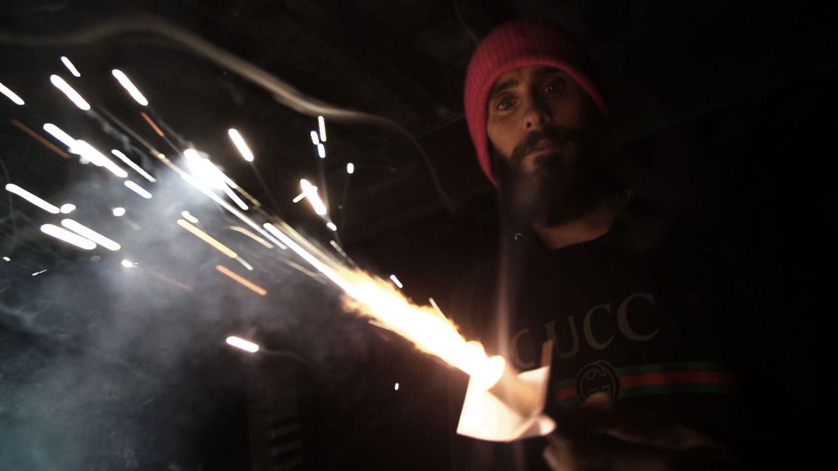 Actor Jared Leto, wearing a red beanie, lights a blow torch in the dark.