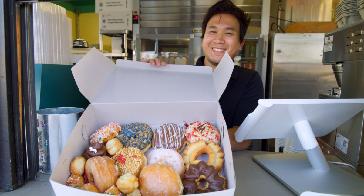 A smiling Cambodian American donut clerk offers up a box full of donuts in Southern California