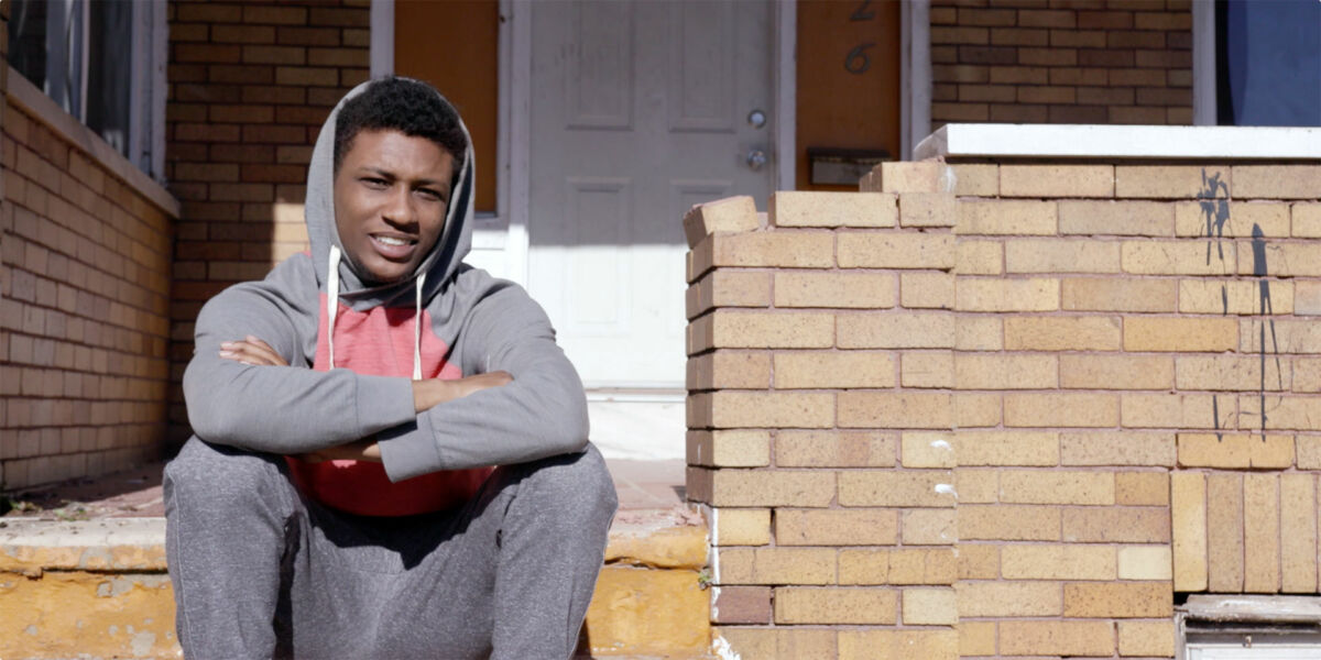 A young man with dark skin tone sits on the stairs of a brick house.