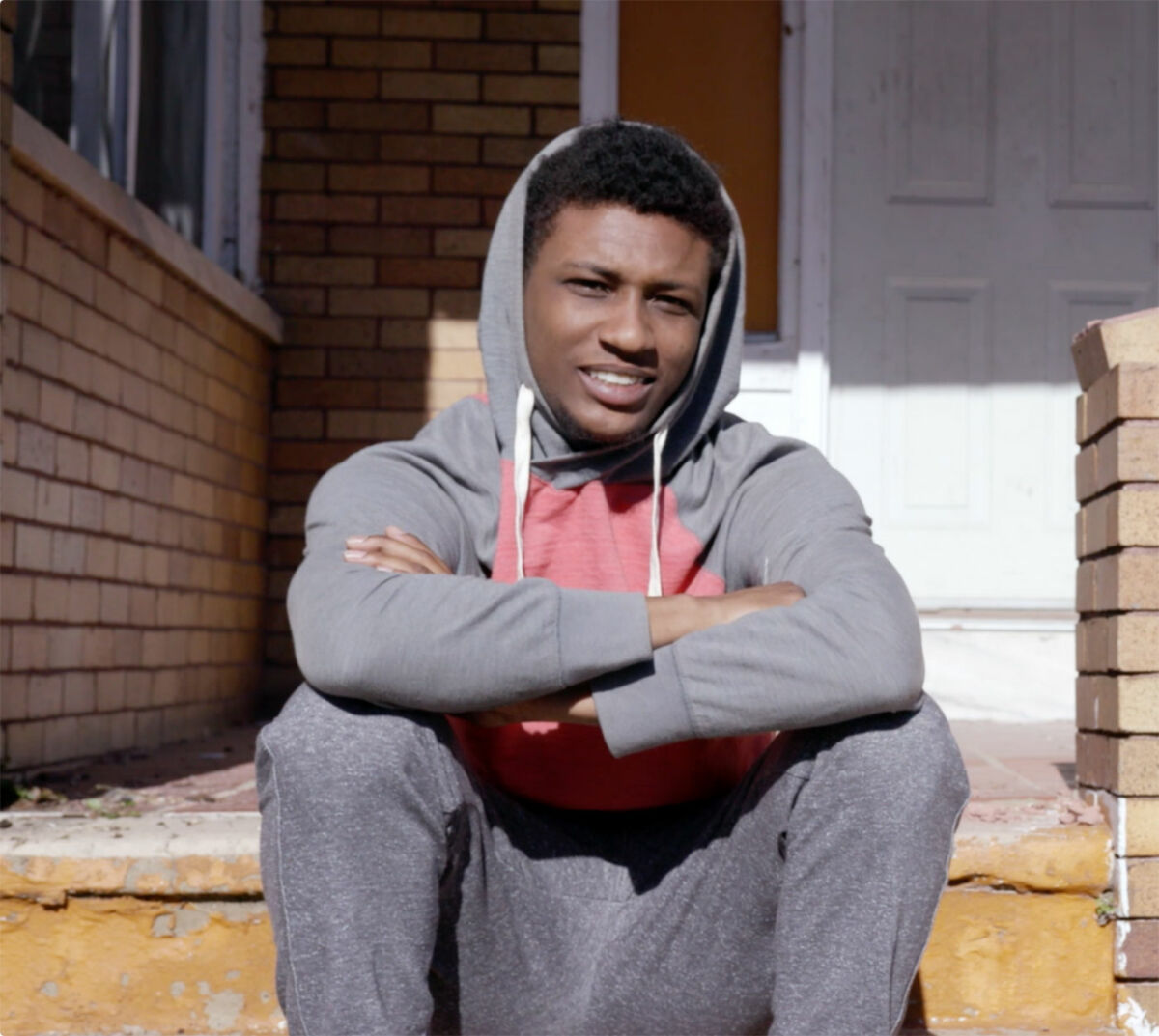 A young man with dark skin tone sits on the stairs of a brick house.