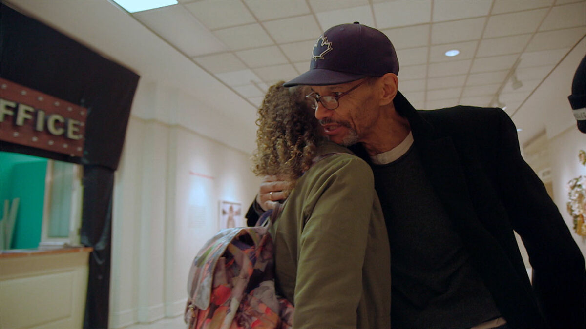 A man with medium-dark skin tone hugs a young woman with curly hair in the hallway of a school.