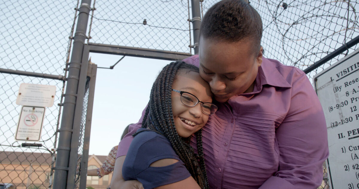 A woman with dark skin tone, wearing a purple shirt, hugs her grinning daughter, a young girl with dark skin tone and long braids.