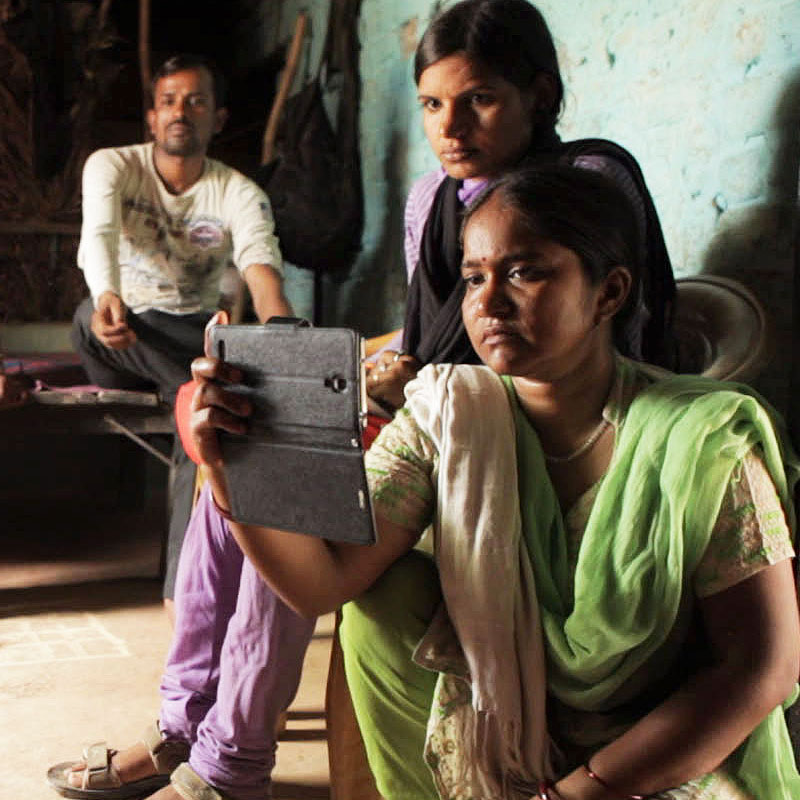 A woman in a green and gold sari videotapes an interview with her smartphone. Another woman looks on behind her.