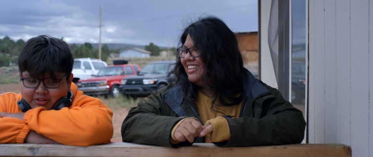 Two Indigenous American teens laugh together outside near a parking lot.