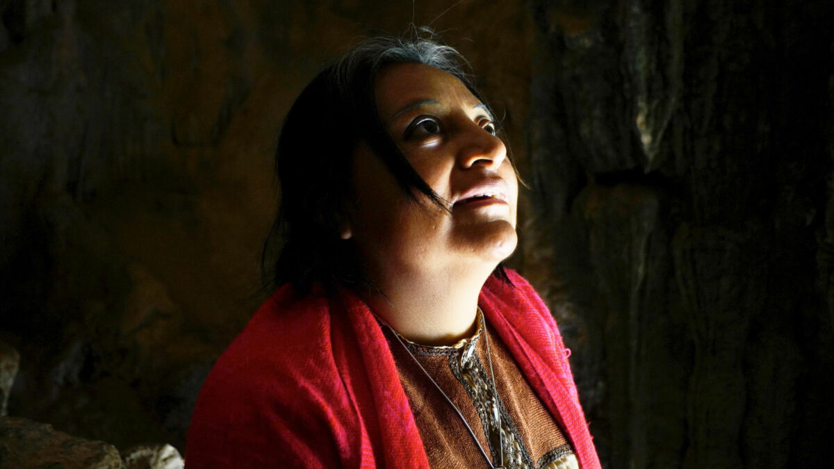 A Mayan healer in a dark room looks up towards the sky with a single source of light shining on her face.