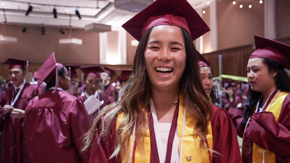 An Asian American student smiles at graduation, wearing a burgundy cap and gown.