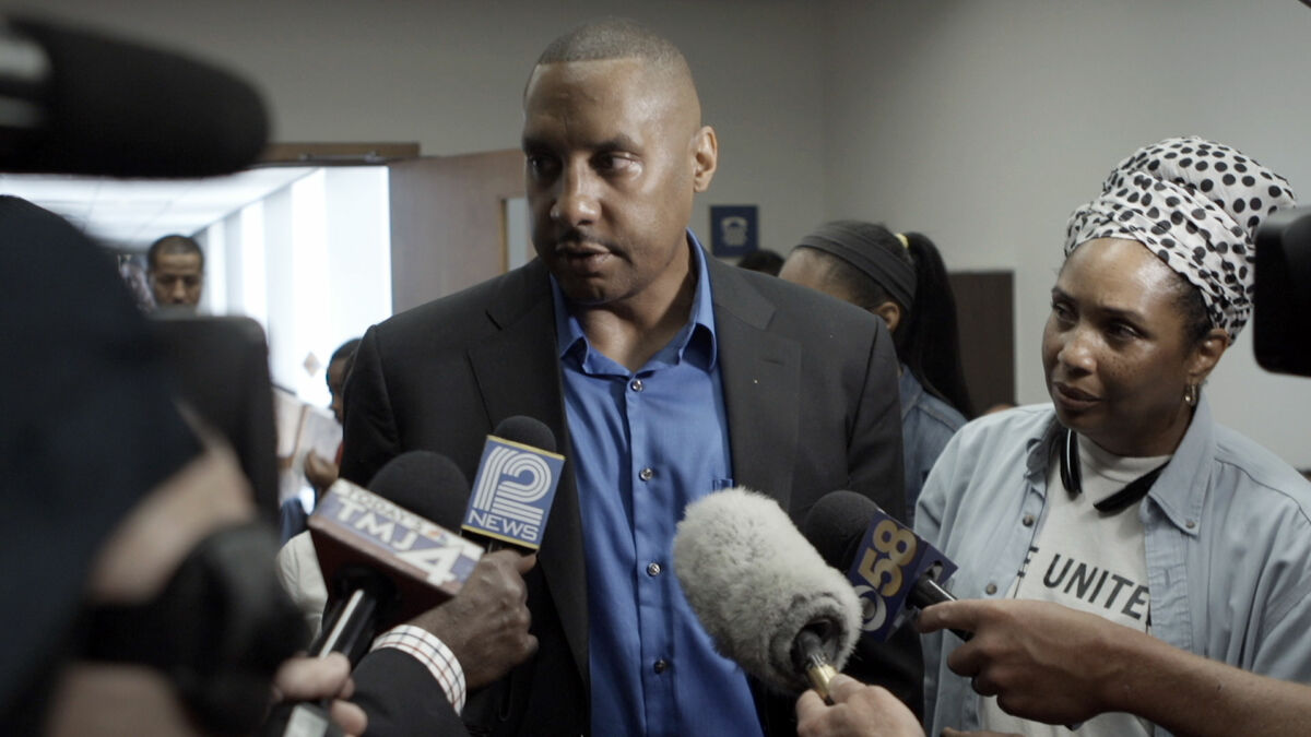 A man in a suit with medium-dark skin tone talks to reporters while a woman with medium-dark skin tone looks on.