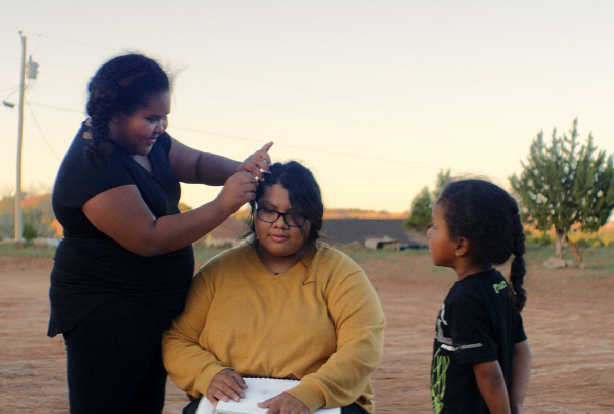 A young Indigenous American girl plays with another's hair while a third looks on outside on the Navajo Nation Reservation.