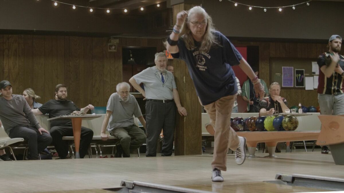 A woman releases a bowling ball down a lane as onlookers wait for the result. String lights adorn the surrounding bowling alley.