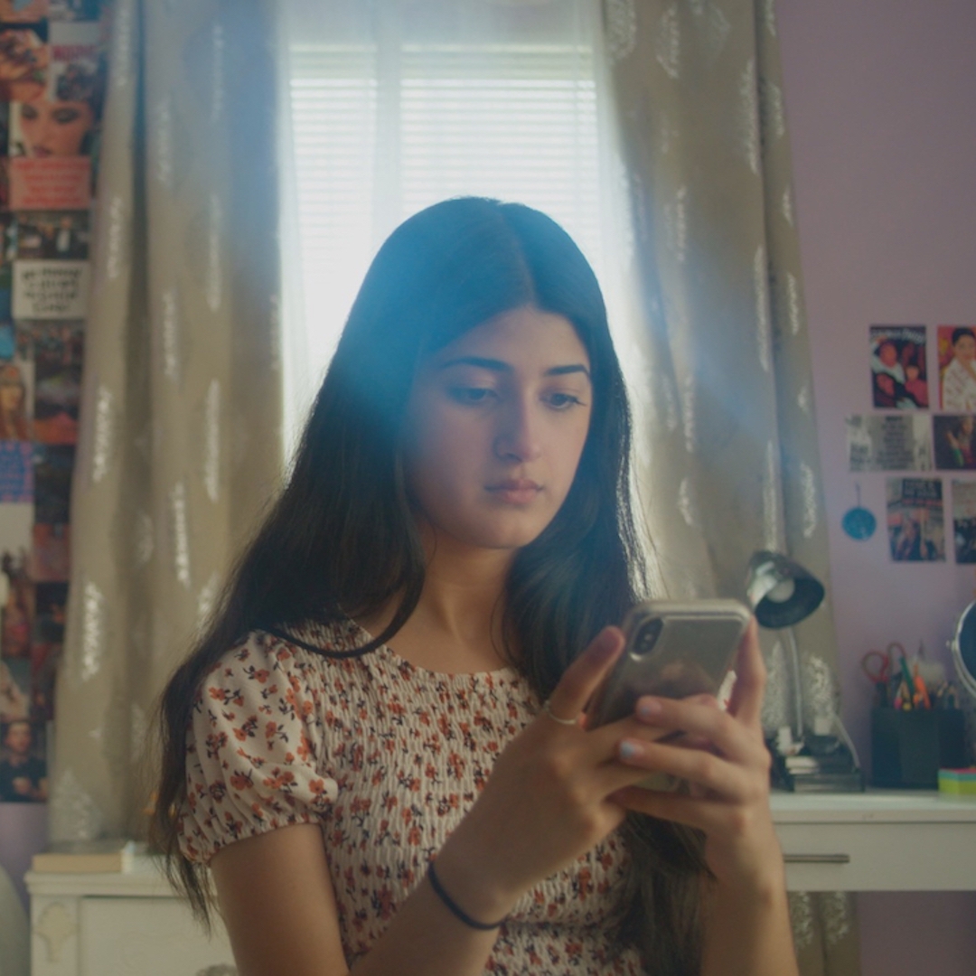 Young woman looks at her phone while in her bedroom with pink walls, surrounded by photographs and magazine cut outs.