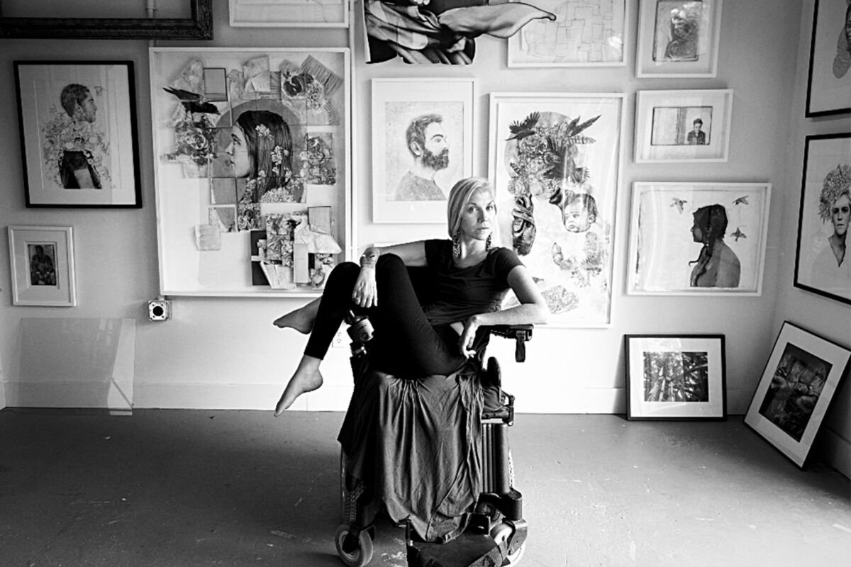 Black and white portrait of a woman posing defiantly in a wheelchair. Surrounding her is a gallery wall of different artwork.