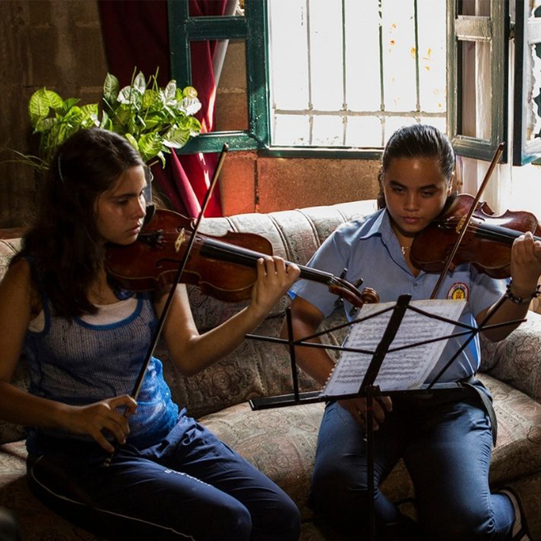 Two girls with medium-light skin tone read sheet music and play the violin while sitting on a floral couch.