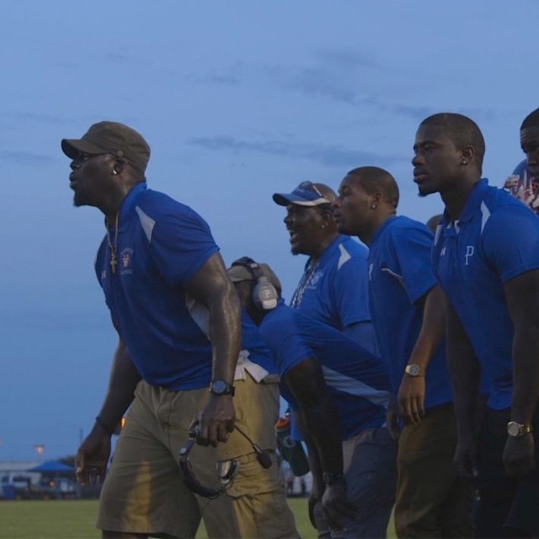 A group of Black coaches and football players sit on the sidelines of a field, energized by the game they are watching.