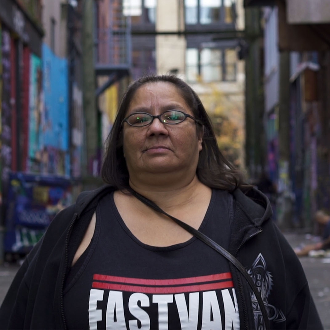 A woman stands facing the camera in an alley covered in murals. She wears glasses and a shirt reading "EASTVAN."