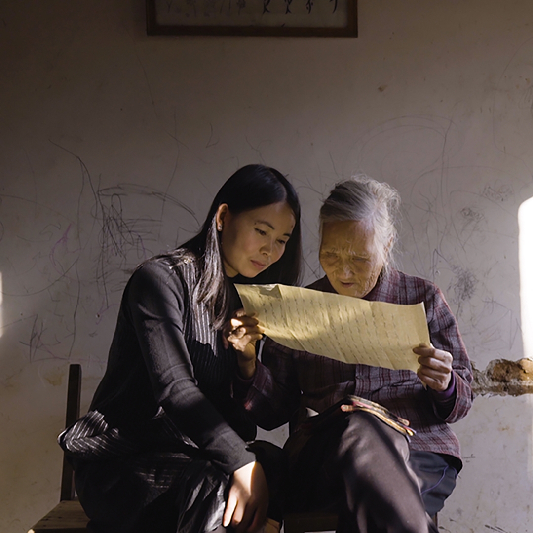 A young woman and an elderly woman reading a letter in a room by the light of a single window.