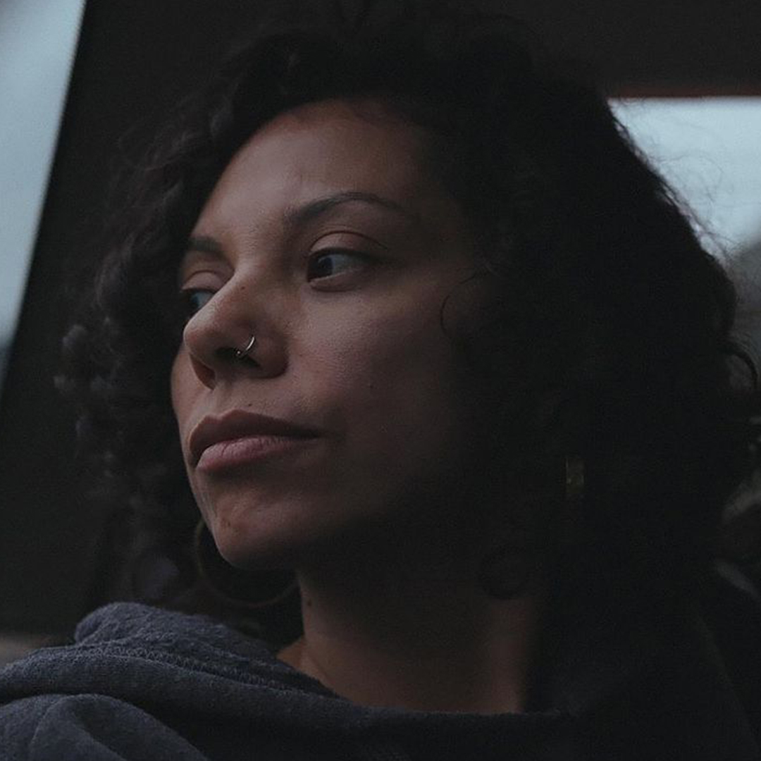 A woman with light skin tone, curly dark hair, and a silver nose ring looks out of a car window.