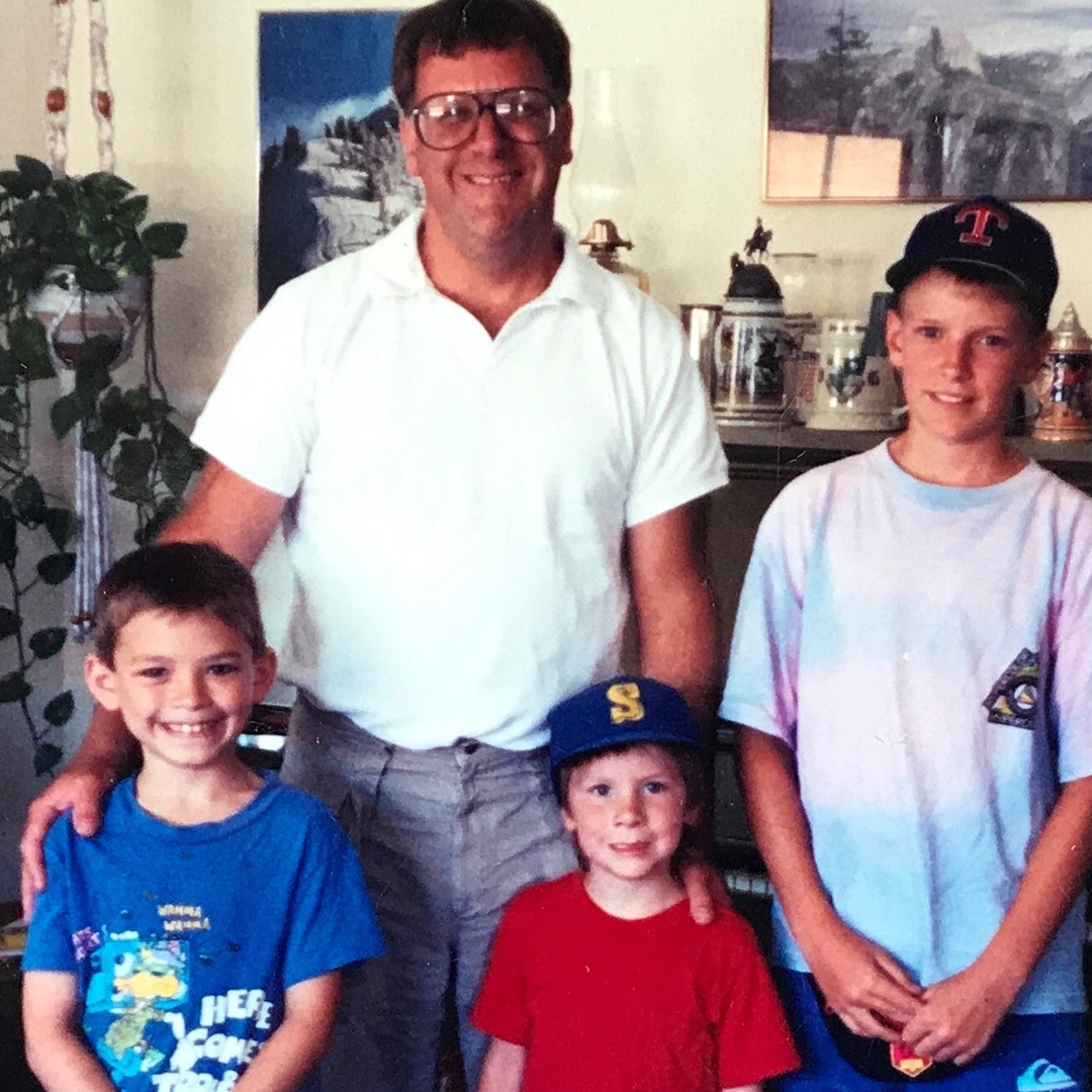 Family photo of a father and three sons in a living room. All are smiling as they pose in front of an upright piano covered in souvenir beer steins and photographs of mountains hung on the wall.