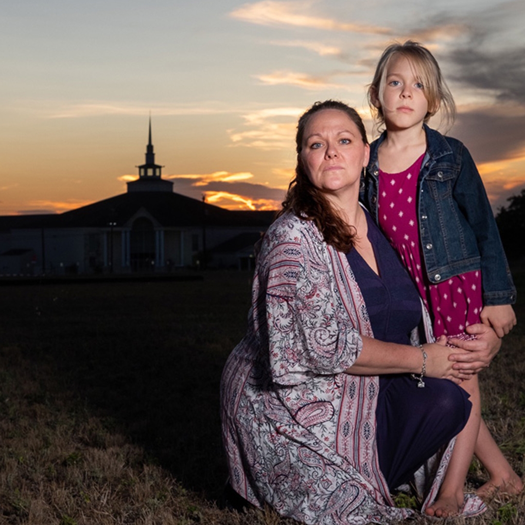 A mother and her daughter, both with light skin tone, pose together in front on a sunset. The steeple of a church is backlit behind them.