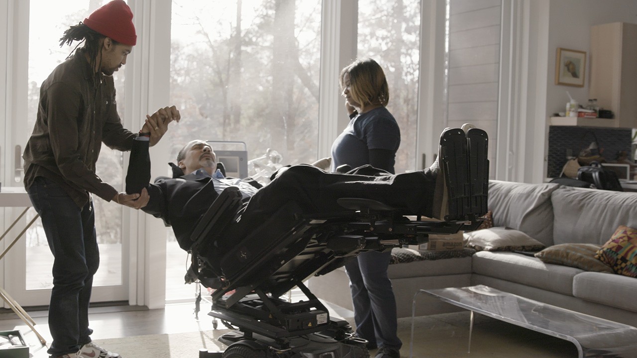 Two people in a bright, light-filled room with tall windows help a man in a wheelchair lean back and stretch his arm.