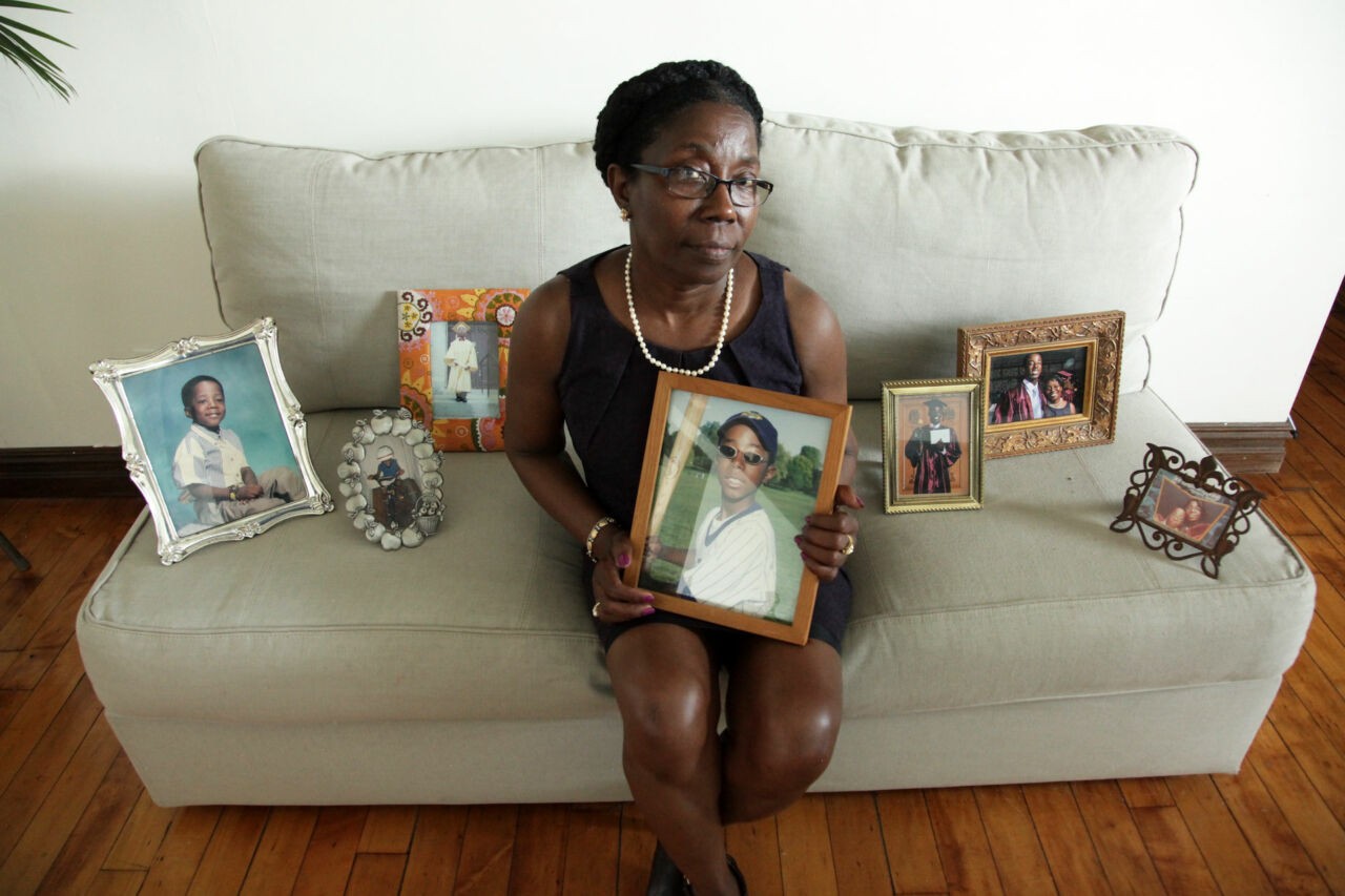 A woman with dark skin tone, wearing a black dress and pearl necklace, sits on a white couch surrounded by photos of her son who has passed due to hazing.