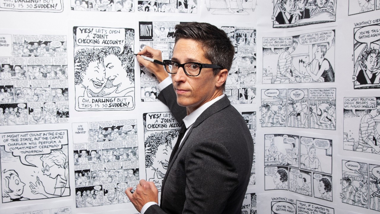 A cartoonist wears a suit in front of a wall of black and white comic strips.