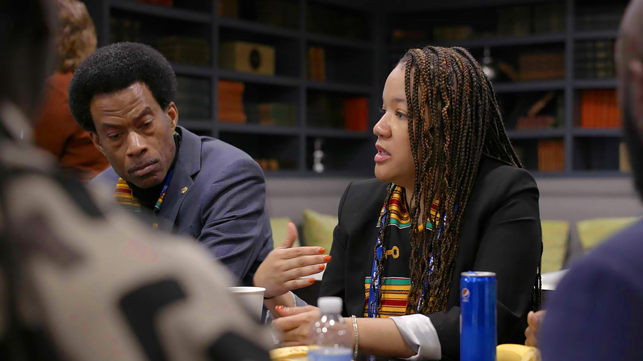 Woman with medium skin tone and long braids sits at a table speaking energetically with others.