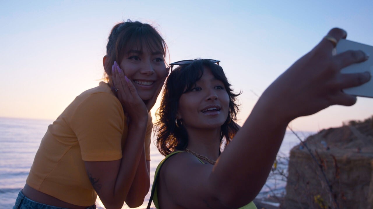 Two young girls, backlit by the sun, take a selfie in front of the ocean.