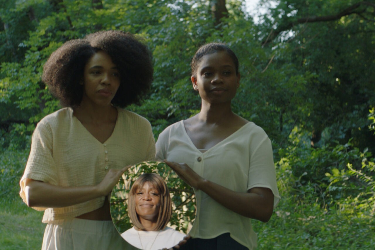 Two women with dark skin tone hold a mirror for a third woman with dark skin tone so she can observe herself. The three women are surrounded by trees.