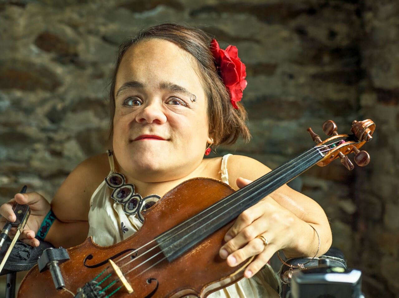 A photo of a violinist with osteogenesis imperfecta holding her instrument. She wears a red flower in her hair and has a eyebrow ring.