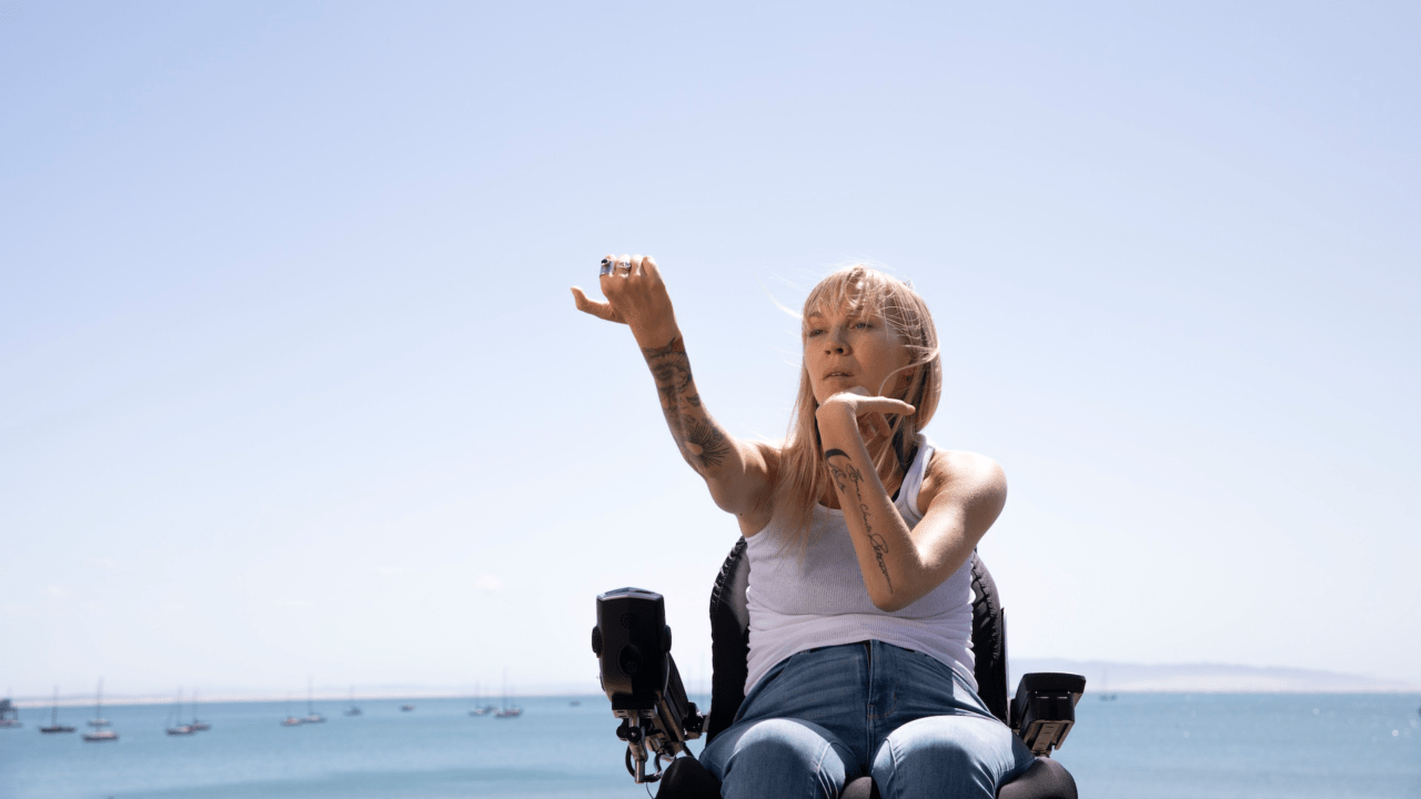 A woman using a wheelchair dances with her arms as she sits near the seaside.