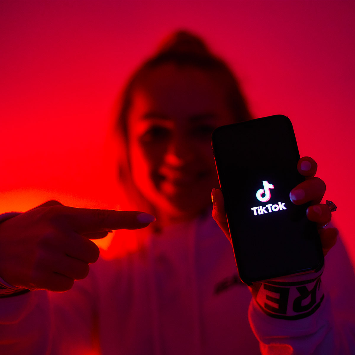 A young girl lit by a red light points to her smartphone screen, which has the TikTok logo on it.