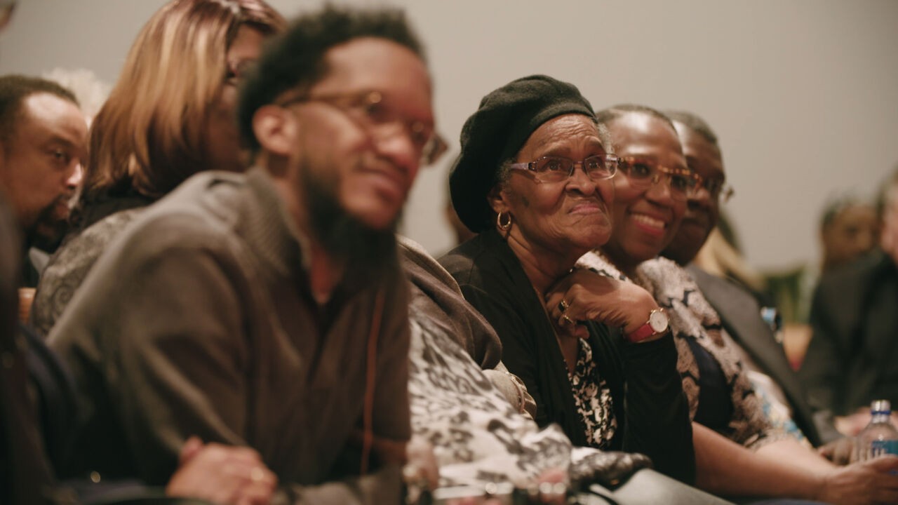 A group of Black men and women sitting in a row, watching someone speak.