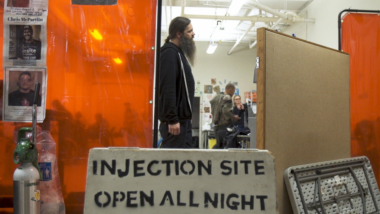 An injection site in Vancouver with orange colored partitions separating different areas. A sign reads "Injection site open all night." A man with a ponytail and long beard stands in the middle of the site looking off camera to the right.