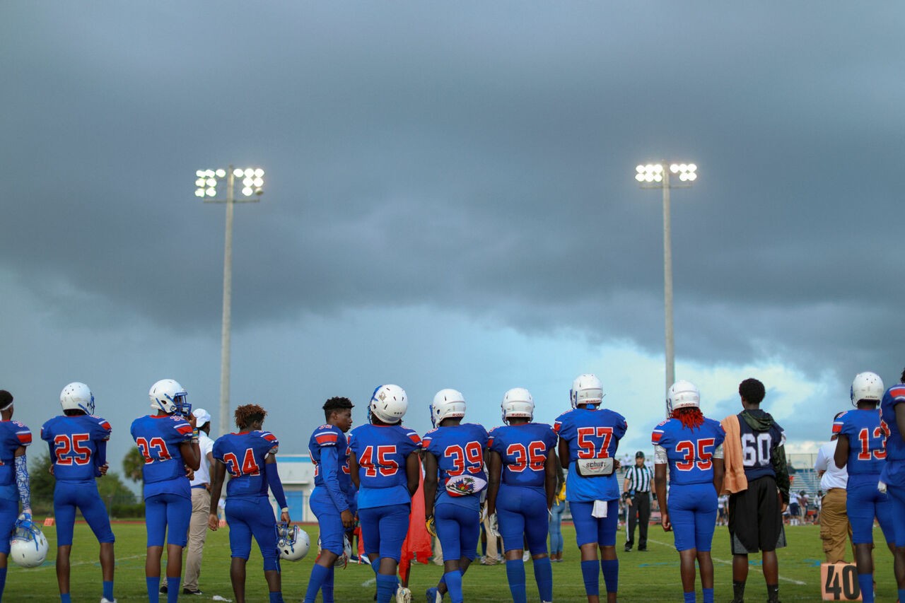 A group of football players stand on the sidelines of a field during a game.