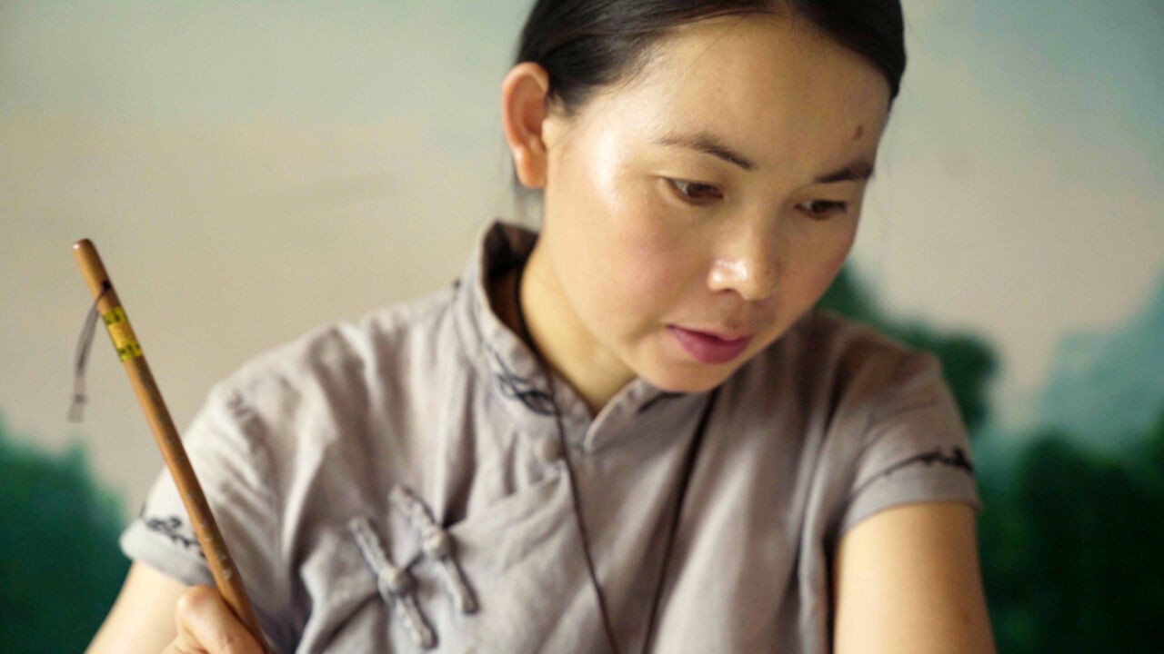 A woman with light skin tone and dark hair wears a qipao and writes with a calligraphy brush.