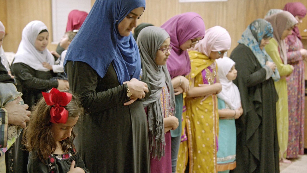 Several women in colorful hijabs bow their heads in prayer inside a wood-paneled Islamic center in Texas.