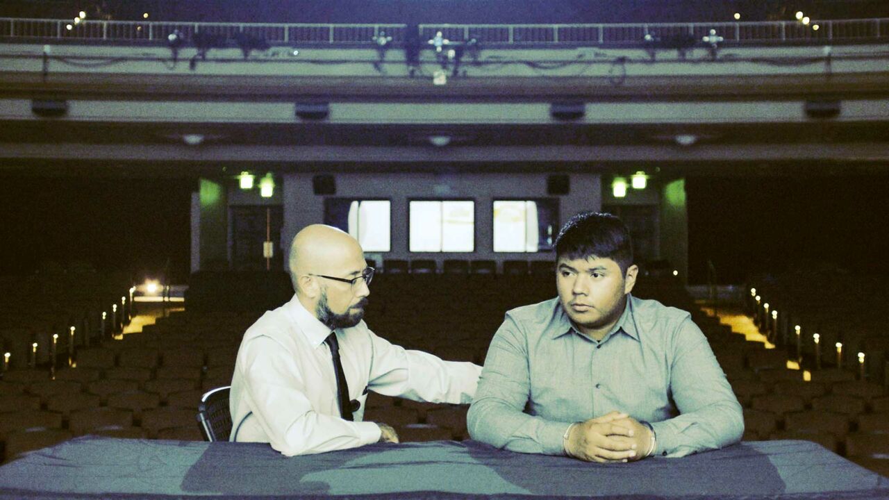 Filmmaker Rodrigo Reyes, a Mexican-American man with medium-light skin tone and glasses sits next to a man with medium skin tone and dark features at a table in an auditorium, recreating a courtroom scene.