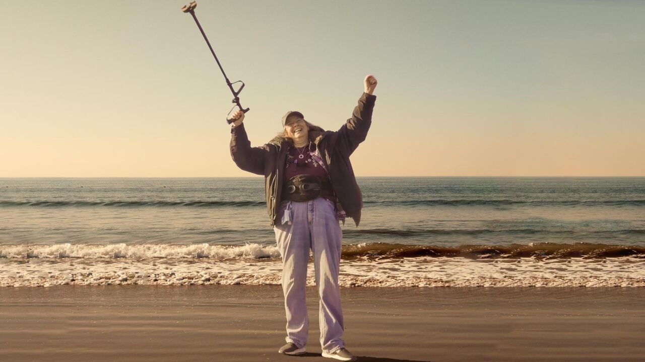 A woman with light skin tone and gray hair stands in front of the ocean. She has raised both of her arms, holding her walking cane up to the sky in a cheer.